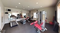 Located at 9295 North Coursier Lane in Spokane, Washington, this single-floor fitness center is thoughtfully designed to support a full range of workout routines. The space features a neutral color palette with warm beige walls, recessed lighting, and durable dark rubber flooring suited for high-traffic athletic use. Multiple windows fitted with wooden blinds allow natural light to fill the room, creating a bright and energizing environment.

The main workout area is well-equipped with a variety of cardio machines, including a treadmill, elliptical, and stationary recumbent bike. Strength training is thoroughly covered with a multi-station cable machine, adjustable weight benches, and a dedicated dumbbell rack stocked with a full range of free weights. Kettlebells are also available for functional training exercises. A large wall-mounted mirror enhances the sense of space and allows users to monitor their form during workouts.

A secondary area near a glass exterior door provides additional space and natural light, housing the free weight section alongside informational boards and reference materials mounted on the wall. A water cooler is conveniently positioned within the facility. The overall layout is organized and functional, offering users everything needed for a comprehensive fitness routine in a clean, well-maintained setting.