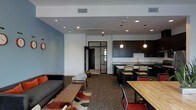 This single-floor space is designed with a contemporary, open-concept layout that seamlessly integrates multiple functional zones into one cohesive environment. The lounge area is anchored by a charcoal sofa, accent chairs, and a low wooden coffee table, set atop a patterned area rug that adds warmth and visual interest to the space. A soft blue accent wall provides a calming backdrop, complementing the neutral white ceilings outfitted with recessed lighting and track lighting for flexible illumination throughout. Adjacent to the lounge, a fully equipped kitchen features dark cabinetry, a light-toned countertop, and a sink, illuminated overhead by a pair of globe pendant lights that lend a stylish, modern touch. A raised kitchen bar with bar stools provides a casual seating option that bridges the kitchen and the wider dining area. The dining zone accommodates a large wooden communal table surrounded by industrial-style chairs with red metal frames, making it well-suited for group gatherings or collaborative work. A glass-paneled door leads to an adjoining room, enhancing the sense of openness and connectivity. The patterned carpet flooring runs throughout, adding texture underfoot and contributing to the space's inviting, functional atmosphere.