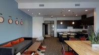 This single-floor space is designed with a contemporary, open-concept layout that seamlessly integrates multiple functional zones into one cohesive environment. The lounge area is anchored by a charcoal sofa, accent chairs, and a low wooden coffee table, set atop a patterned area rug that adds warmth and visual interest to the space. A soft blue accent wall provides a calming backdrop, complementing the neutral white ceilings outfitted with recessed lighting and track lighting for flexible illumination throughout. Adjacent to the lounge, a fully equipped kitchen features dark cabinetry, a light-toned countertop, and a sink, illuminated overhead by a pair of globe pendant lights that lend a stylish, modern touch. A raised kitchen bar with bar stools provides a casual seating option that bridges the kitchen and the wider dining area. The dining zone accommodates a large wooden communal table surrounded by industrial-style chairs with red metal frames, making it well-suited for group gatherings or collaborative work. A glass-paneled door leads to an adjoining room, enhancing the sense of openness and connectivity. The patterned carpet flooring runs throughout, adding texture underfoot and contributing to the space's inviting, functional atmosphere.