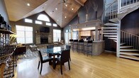 This single-floor space showcases a dramatic open-concept layout unified by soaring vaulted ceilings clad in warm wood paneling, which lend the interior a sense of grandeur and airiness. A ceiling fan is centrally mounted overhead, complementing recessed lighting throughout. Natural light pours in through an arched transom window and multiple flanking windows above the living area, brightening the space considerably.

The living area is anchored by a floor-to-ceiling stacked stone fireplace with a mounted flat-screen television above, creating a cozy yet refined focal point. Adjacent to it, a glass-top round dining table with dark leather chairs sits atop a patterned area rug over rich hardwood flooring.

The kitchen area features a substantial bar-height island with sleek black bar stools, light wood cabinetry, and a decorative mantel-style surround that adds architectural character. A spiral staircase along one wall adds a stylish vertical element. Deep blue-gray accent walls contrast elegantly with the warm wood tones, lending the overall space a sophisticated, contemporary ambiance.