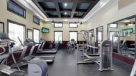 This fitness center offers a thoughtfully designed workout space that combines functionality with refined architectural detail. The room is defined by a striking coffered ceiling with dark-framed recessed panels, recessed lighting, and stylish pendant light fixtures that lend a polished, upscale ambiance uncommon in typical gym settings.

Natural light flows in through a series of windows positioned along the upper and lower portions of the walls, creating a bright and energizing environment. The walls feature a two-tone treatment with a deep burgundy lower section and a neutral cream upper half, adding visual depth to the space.

The flooring consists of durable dark rubber matting, well-suited for heavy equipment and high-traffic use. Cardio equipment, including multiple treadmills and stationary bikes by Precor, lines one side of the room, while cable and resistance machines are arranged to allow comfortable circuit-style training. A wall-mounted flat-screen television is positioned for convenient viewing during workouts. Mirrors along one wall enhance the sense of space and allow users to monitor their form. The overall atmosphere is clean, organized, and inviting.