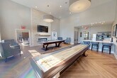 Located at 1100 Jericho Turnpike in Commack, NY, this single-floor community clubhouse offers a generously sized, thoughtfully designed gathering space. The layout encompasses a living room, a dedicated dining room, and a kitchen/living area, all unified by warm herringbone-patterned hardwood flooring and a neutral, light-toned color palette.

The living room serves as the centerpiece, featuring soaring double-height ceilings, a striking floor-to-ceiling stacked stone fireplace with a wall-mounted television above, and a pool and shuffleboard table for recreation. Plush blue armchairs add a welcoming touch, while an abundance of oversized windows and glass doors flood the room with natural light and offer views of the surrounding outdoor areas.

The dining room is an elegant space furnished with multiple dining tables and upholstered chairs in soft blue tones. Large pendant drum lights illuminate the room, complemented by natural light streaming through tall windows. A built-in buffet with recessed lighting anchors one wall, adding a refined finishing touch.

The kitchen is efficiently designed with stainless steel appliances, gray cabinetry, white subway tile backsplash, and clean countertops. Glass-paneled interior doors connect the spaces with transparency and style. Curated black-and-white artwork displayed throughout ties the aesthetic together, creating a cohesive, modern, and inviting environment.