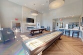 Located at 1100 Jericho Turnpike in Commack, NY, this single-floor community clubhouse offers a generously sized, thoughtfully designed gathering space. The layout encompasses a living room, a dedicated dining room, and a kitchen/living area, all unified by warm herringbone-patterned hardwood flooring and a neutral, light-toned color palette.

The living room serves as the centerpiece, featuring soaring double-height ceilings, a striking floor-to-ceiling stacked stone fireplace with a wall-mounted television above, and a pool and shuffleboard table for recreation. Plush blue armchairs add a welcoming touch, while an abundance of oversized windows and glass doors flood the room with natural light and offer views of the surrounding outdoor areas.

The dining room is an elegant space furnished with multiple dining tables and upholstered chairs in soft blue tones. Large pendant drum lights illuminate the room, complemented by natural light streaming through tall windows. A built-in buffet with recessed lighting anchors one wall, adding a refined finishing touch.

The kitchen is efficiently designed with stainless steel appliances, gray cabinetry, white subway tile backsplash, and clean countertops. Glass-paneled interior doors connect the spaces with transparency and style. Curated black-and-white artwork displayed throughout ties the aesthetic together, creating a cohesive, modern, and inviting environment.