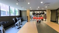 This single-level fitness center is thoughtfully designed to accommodate a wide range of workout routines. The space features a combination of rubber and hard flooring zones, clearly delineating areas for different types of exercise. Recessed ceiling lighting provides bright, even illumination throughout, while a window along one wall allows natural light to filter in, offering a view of the surrounding streetscape.

The cardio area is equipped with stationary cycling bikes, ideal for high-intensity interval or endurance training. A dedicated functional training zone houses an organized rack of medicine balls and stability balls in varying sizes, supporting strength, balance, and core conditioning exercises. A large mirrored wall expands the visual depth of the space and allows users to monitor their form during workouts.

Beyond the mirror partition, additional equipment is visible, suggesting a well-stocked free weights and strength training section. A column with an adjacent storage rack for weighted balls adds to the organized layout. The overall design combines a dark, contemporary aesthetic with practical fitness infrastructure, creating a focused and motivating training environment.