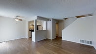 This two-story home offers a thoughtfully laid-out interior designed for comfortable everyday living. The main level features an open-concept living area finished with warm hardwood-style flooring that extends throughout the space, creating a cohesive and inviting atmosphere. A textured ceiling adds visual interest overhead, while neutral gray walls provide a clean, modern backdrop.

The kitchen is accessible via a pass-through opening framed by a half-wall, allowing for a connected feel between the cooking and living areas. It is equipped with white cabinetry, stainless steel appliances including a refrigerator and range, and a functional under-cabinet range hood. Recessed lighting illuminates the kitchen workspace efficiently.

From the main level, a staircase with white balusters leads to the second floor, offering additional living space above. A hallway visible from the living area suggests further rooms and storage options on the main level as well. The overall design is practical and well-organized, with clean lines and a neutral palette that makes the space feel bright and versatile. The layout is well-suited for those seeking a functional multi-level home with clearly defined yet openly connected living spaces.