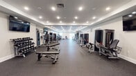 This fitness center at 1 E Pearl St in Burlington, New Jersey, offers a spacious, single-floor layout designed to accommodate a wide range of workout routines. The facility is thoughtfully organized to maximize floor space, with durable rubber flooring throughout that provides both safety and comfort during exercise.

The strength training area is extensively equipped with Atlantis-brand machines, including multi-functional cable systems, plate-loaded equipment, and a full dumbbell rack with adjustable benches. A dedicated cardio zone features multiple NordicTrack treadmills positioned near natural light sources, along with elliptical machines and additional cardio equipment. Wall-mounted flat-screen TVs are positioned throughout for an engaging workout experience.

The interior design features a clean, modern aesthetic with tray ceilings, recessed lighting, and neutral-toned walls that create a bright and open atmosphere. Large windows and sliding glass doors in the cardio area allow natural light to fill the space, contributing to an energizing environment. A water station is conveniently accessible within the facility, and double doors provide easy entry and exit. The overall layout flows efficiently, offering ample open floor space between equipment stations to ensure a comfortable and functional training experience.