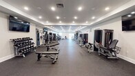 This fitness center at 1 E Pearl St in Burlington, New Jersey, offers a spacious, single-floor layout designed to accommodate a wide range of workout routines. The facility is thoughtfully organized to maximize floor space, with durable rubber flooring throughout that provides both safety and comfort during exercise.

The strength training area is extensively equipped with Atlantis-brand machines, including multi-functional cable systems, plate-loaded equipment, and a full dumbbell rack with adjustable benches. A dedicated cardio zone features multiple NordicTrack treadmills positioned near natural light sources, along with elliptical machines and additional cardio equipment. Wall-mounted flat-screen TVs are positioned throughout for an engaging workout experience.

The interior design features a clean, modern aesthetic with tray ceilings, recessed lighting, and neutral-toned walls that create a bright and open atmosphere. Large windows and sliding glass doors in the cardio area allow natural light to fill the space, contributing to an energizing environment. A water station is conveniently accessible within the facility, and double doors provide easy entry and exit. The overall layout flows efficiently, offering ample open floor space between equipment stations to ensure a comfortable and functional training experience.