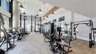 Located at The Townsend in San Diego, CA, this two-story fitness center offers a thoughtfully designed space for health and wellness. The main floor welcomes visitors through a sleek, modern lobby featuring polished concrete floors, cylindrical columns, and expansive floor-to-ceiling windows that flood the entry with natural light and frame views of the surrounding lush greenery. A built-in service station with wood cabinetry, a mini refrigerator, and a mounted display screen adds a functional touch near the entrance. The primary exercise room is an impressive, double-height space anchored by soaring ceilings with exposed wood beam accents, bold pendant lighting, and a full wall of windows overlooking the outdoors. It is equipped with treadmills, cable machines, free weights, a rowing machine, and multi-use training stations, all set atop patterned carpet and concrete flooring. Full-length mirrors maximize the sense of space throughout. A striking staircase with black metal railings leads to the upper level, where light-toned hardwood floors and recessed lighting define a secondary open exercise area. An outdoor terrace extends the fitness experience further, featuring a freestanding training rig with a punching bag amid views of palm trees and the San Diego skyline.