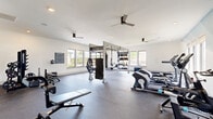 This spacious fitness center offers a comprehensive workout environment spread across a single open floor. The space is designed with a clean, modern aesthetic, featuring crisp white ceilings, recessed lighting, and multiple ceiling fans that ensure comfortable air circulation throughout. Large windows line the walls, flooding the interior with abundant natural light and providing views of the surrounding outdoors.

The facility is divided into distinct workout zones. A dedicated cardio area houses elliptical machines, stationary bikes, and treadmills, complemented by a wall-mounted flat-screen TV. A strength training section features a multi-station functional rig with TRX suspension trainers, battle ropes, and a heavy punching bag, catering to dynamic and functional fitness routines. A free weights area includes a full dumbbell rack and adjustable benches, while a rowing machine offers an additional cardio option.

Full-length mirrors are strategically placed to enhance the sense of space and allow users to monitor their form. Durable rubber flooring covers the exercise areas, providing safety and impact absorption. A fitness rules board is posted on the wall, emphasizing a well-managed facility. The overall ambiance is bright, open, and professional, making this fitness center a well-rounded space suitable for a wide range of training styles and fitness levels.