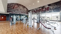 Located at 301 Church Ave in College Station, TX, this fitness center offers a generously sized, single-floor layout comprising two dedicated exercise rooms. The space is defined by its energetic, sports-inspired aesthetic, with bold maroon-and-grey motivational murals spanning the walls and ceiling — featuring powerful phrases like "Make It Happen," "Discipline," and "Perseverance" alongside dynamic athletic imagery that sets an inspiring tone throughout.

The main exercise room is expansive and open, with warm hardwood-style flooring, soaring arched ceilings, and recessed lighting. It is outfitted with a comprehensive selection of strength training machines, including cable systems, multi-use weight stations, and Cybex Prestige equipment, as well as free weight racks lined with dumbbells and adjustable benches. Floor-to-ceiling windows line the perimeter, flooding the space with natural light and offering views of the surrounding area.

The second exercise room features a focused cardio zone, housing treadmills, elliptical trainers, stair climbers, and stationary bikes, all positioned along the large windows to maximize natural light. Full-length mirrors, medicine balls, and additional accessories round out the space. Together, both rooms provide a well-equipped, motivating environment suited for a complete workout experience.