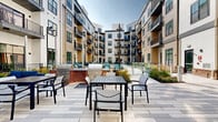Located at 311 Karl Linn Drive in North Chesterfield, Virginia, this modern residential community is defined by its thoughtfully designed outdoor amenity spaces and contemporary architectural character. The multi-story building wraps around a central courtyard, creating an enclosed, resort-like atmosphere that feels both private and expansive.

At the heart of the community is a large, crystal-clear swimming pool surrounded by a generous sun deck lined with lounge chairs, perfect for relaxing outdoors. Glass pool fencing adds a sleek, open feel while maintaining safety and sightlines throughout the courtyard. Adjacent to the pool, a dedicated outdoor grilling and dining area features stainless steel grills set atop a brick base, accompanied by a patio dining table and chairs — ideal for al fresco gatherings.

The building's exterior showcases a blend of warm neutral siding and classic brick detailing, accented by bold dark-framed windows and private balconies on upper levels. Landscaped walkways wind along the building's perimeter, featuring ornamental grasses, shrubs, and seasonal plantings that add greenery and texture to the hardscaped surroundings. Pole-mounted lantern-style lighting enhances the ambiance after dark. The overall environment is clean, contemporary, and inviting, offering residents a well-appointed outdoor living experience.