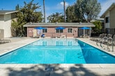 Located at 100 S Seranado St in Orange, California, this single-story residential community offers a welcoming outdoor environment designed for comfort and leisure. The property is defined by its generously sized swimming pool, which serves as the centerpiece of the communal outdoor space. The pool area is surrounded by a wide concrete deck lined with lounge chairs and shaded by large, mature trees that provide a sense of natural enclosure and privacy.

The buildings feature a classic Southern California architectural style, with neutral stucco and wood-paneled exteriors accented by pitched rooflines. Lush greenery, including flowering shrubs and established trees, is integrated throughout the grounds, contributing to a park-like setting. A secured gated entry separates portions of the outdoor area, adding an element of privacy and safety to the community.

Shaded seating areas with stone tables and umbrella-covered spots offer additional outdoor gathering spaces adjacent to the pool. The layout of the property, as seen from an aerial perspective, reveals a thoughtfully arranged courtyard-style design with buildings enclosing the central amenity area. Under the clear blue skies typical of Orange County, the space exudes a bright, relaxed ambiance well-suited to the Southern California lifestyle.