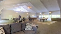 This single-floor recreational space is defined by its striking vaulted ceiling with exposed white beams that rise dramatically overhead, creating an open and airy atmosphere throughout the room. Multiple ceiling fans and pendant light fixtures are suspended from the ceiling, ensuring the space remains well-lit and comfortable. Large windows along the perimeter walls allow generous amounts of natural light to flood the interior, enhancing the bright and welcoming ambiance.

The layout is thoughtfully divided into distinct activity zones. A lounge area at the front features a comfortable gray sofa with accent pillows and a side table, ideal for casual gathering and relaxation. Moving further into the space, a dining or card-playing area is furnished with multiple tables and chairs, suitable for group activities. Toward the rear, the space transitions into a game room area equipped with what appears to be pool tables, providing entertainment options for residents or guests.

Bookshelves and storage units line portions of the walls, adding functional elements to the space. The neutral color palette of white walls and light carpeting contributes to the clean, open feel of this versatile communal facility.