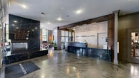 The Cliffs Lofts, located at 100 Paterson Plank Rd in Jersey City, NJ, presents a generously sized lobby and amenity floor that masterfully blends industrial heritage with modern sophistication. Polished, marbled concrete floors flow seamlessly throughout, reflecting light and anchoring the space's contemporary aesthetic.

The entrance foyer welcomes visitors with exposed brick columns, glass-paneled doors, and recessed lighting, setting a refined tone from the moment of arrival. The front desk area features a dramatic black marble reception counter beneath the prominently displayed "The Cliffs Lofts" signage, framed by rich wood accents and steel-framed glass partitions.

The main lounge area exudes an urban loft atmosphere with soaring ceilings, exposed brick walls, large arched multi-pane windows flooding the space with natural light, and curated furnishings including leather sofas and sculptural accent chairs. A dedicated meeting or dining room offers a substantial wood table, orange accent chairs, a beverage station, and bold East Asian brushstroke artwork. A media lounge with dark walls, a large flat-screen TV, and a sleek credenza provides a more intimate retreat. A children's playroom features soft carpet, tall arched windows with garden views, whimsical wall murals, and an array of toys, making the space welcoming for all residents.