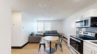 Located at 221 E State St in West Lafayette, Indiana, this one-bedroom, one-bathroom unit offers a well-organized and functional layout across a single floor. The open-concept living and kitchen area serves as the heart of the space, where light wood flooring and white walls create a clean, bright atmosphere. The kitchen is outfitted with stainless steel appliances — including a refrigerator, electric range, microwave, and dishwasher — complemented by white shaker-style cabinetry and a gray subway tile backsplash that adds a modern touch. A round dining table with metal chairs sits adjacent to the kitchen, ideal for casual meals. The living area provides comfortable seating and is brightened by windows that invite natural light and outdoor views. The bedroom is a cozy, private retreat with a window overlooking the surroundings and space for a desk, making it well-suited for work or study. Two closets offer practical storage throughout the unit. The bathroom features a combined tub and shower with clean white finishes and wood-look flooring consistent with the rest of the space. Textured ceilings and dark baseboards add subtle character throughout this efficiently designed unit.