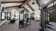 This expansive fitness center is a thoughtfully designed facility that offers a comprehensive workout experience across multiple distinct zones. The space is defined by its striking architectural character — soaring vaulted ceilings adorned with rich, dark-stained exposed wood beams run throughout, creating an open and airy atmosphere complemented by ceiling fans and recessed lighting.

The main strength and cardio area features plush gray carpet flooring and is outfitted with a wide array of professional-grade equipment, including cable machines, multi-functional strength stations, benches, a squat rack, treadmills, ellipticals, and a punching bag. Large black-framed windows line the walls, inviting generous natural light and offering views to the outdoors.

A dedicated studio room transitions seamlessly from the main floor, distinguished by bold navy patterned accent walls, light wood flooring, and floor-to-ceiling mirrors. This area is equipped with Matrix cycling bikes, kettlebells, medicine balls, and a well-organized weight rack — ideal for group or individual training sessions.

A separate free weights nook provides a focused environment with a full dumbbell rack and mirrored walls. An entry corridor features a wall-mounted TV and connects the zones with ease. Throughout, the design balances high-end finishes with functional, performance-focused equipment for a premium fitness experience.