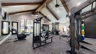 This expansive fitness center is a thoughtfully designed facility that offers a comprehensive workout experience across multiple distinct zones. The space is defined by its striking architectural character — soaring vaulted ceilings adorned with rich, dark-stained exposed wood beams run throughout, creating an open and airy atmosphere complemented by ceiling fans and recessed lighting.

The main strength and cardio area features plush gray carpet flooring and is outfitted with a wide array of professional-grade equipment, including cable machines, multi-functional strength stations, benches, a squat rack, treadmills, ellipticals, and a punching bag. Large black-framed windows line the walls, inviting generous natural light and offering views to the outdoors.

A dedicated studio room transitions seamlessly from the main floor, distinguished by bold navy patterned accent walls, light wood flooring, and floor-to-ceiling mirrors. This area is equipped with Matrix cycling bikes, kettlebells, medicine balls, and a well-organized weight rack — ideal for group or individual training sessions.

A separate free weights nook provides a focused environment with a full dumbbell rack and mirrored walls. An entry corridor features a wall-mounted TV and connects the zones with ease. Throughout, the design balances high-end finishes with functional, performance-focused equipment for a premium fitness experience.