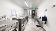 Located at 82 Warren Rd in Toronto's Forest Hill neighbourhood, this shared laundry facility is a well-organized, single-room utility space designed for efficient communal use. The room is lined with a row of commercial-grade LG Direct Drive front-loading washers and dryers finished in stainless steel, providing ample laundry capacity for residents. Two stainless steel folding tables run along the opposite wall, offering generous surface area for sorting and folding laundry. A digital payment kiosk is conveniently positioned near the entrance, enabling cashless transactions. The bright white walls and flush fluorescent ceiling lighting create a clean, well-lit environment throughout. The floor features a distinctive light-toned herringbone-patterned vinyl plank, adding a modern touch to the functional space. A window along one wall admits natural light, while an emergency exit door with an alarm provides additional egress. Informational postings are displayed on the walls, offering usage instructions and building notices. A water filtration unit is tucked near the entry door, and all plumbing connections are neatly recessed into the wall. The space is equipped with a fire extinguisher and smoke detector, reflecting a strong commitment to safety standards.