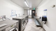 Located at 82 Warren Rd in Toronto's Forest Hill neighbourhood, this shared laundry facility is a well-organized, single-room utility space designed for efficient communal use. The room is lined with a row of commercial-grade LG Direct Drive front-loading washers and dryers finished in stainless steel, providing ample laundry capacity for residents. Two stainless steel folding tables run along the opposite wall, offering generous surface area for sorting and folding laundry. A digital payment kiosk is conveniently positioned near the entrance, enabling cashless transactions. The bright white walls and flush fluorescent ceiling lighting create a clean, well-lit environment throughout. The floor features a distinctive light-toned herringbone-patterned vinyl plank, adding a modern touch to the functional space. A window along one wall admits natural light, while an emergency exit door with an alarm provides additional egress. Informational postings are displayed on the walls, offering usage instructions and building notices. A water filtration unit is tucked near the entry door, and all plumbing connections are neatly recessed into the wall. The space is equipped with a fire extinguisher and smoke detector, reflecting a strong commitment to safety standards.