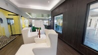 This single-story clubhouse, known as The Ivy, is a thoughtfully designed communal space that blends bold aesthetics with practical amenities. The most striking architectural feature is the large, photo-realistic mural of leafy tree canopies printed across a tray ceiling, creating an immersive, nature-inspired atmosphere throughout the main lounge area. Recessed lighting and subtle LED accents complement the mural, enhancing the overall ambiance.

The main lounge area is generously proportioned, featuring multiple distinct seating zones furnished with crisp white wingback chairs, tufted cream sofas, and vibrant teal and yellow accent seating. A glass-enclosed private lounge with a bold yellow sofa and patterned flooring provides a semi-secluded gathering space. Dark hardwood-style flooring runs throughout, contrasting effectively with the light furnishings and bright color palette.

A dedicated business center lines one wall, equipped with multiple computer workstations and bar-height seating. Adjacent to this is a dark-paneled bar or refreshment counter with decorative sunburst mirrors and stool seating. Dark wood accent walls with large windows offer views to an outdoor patio area, allowing ample natural light to filter in. A bold green entry door adds a further pop of color to this cohesive, design-forward space.