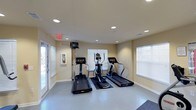 Located at 1749 Dobbins Dr in Chapel Hill, NC, this fitness center offers a well-appointed, single-floor space designed for effective cardiovascular workouts. The room is outfitted with multiple treadmills and an elliptical machine, all positioned on protective rubber floor mats atop a durable tiled floor. Recessed lighting in the smooth ceiling provides consistent, even illumination throughout the space, complemented by several windows fitted with horizontal blinds that allow ample natural light to filter in. The walls are finished in a neutral, light tone that contributes to a clean and open atmosphere. A wall-mounted television is positioned for convenient viewing during workouts. Safety is addressed with a clearly marked exit sign and a mounted fire extinguisher near the entrance. A glass-paneled door offers easy access and an additional source of natural light. The overall layout is thoughtfully organized to maximize usable floor space while maintaining an uncluttered, welcoming environment suitable for focused exercise.