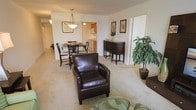 This single-floor residence is designed with an open-concept layout that seamlessly connects the living, dining, and kitchen areas, creating a cohesive and functional flow throughout the space. The living area features plush carpeted flooring in a neutral beige tone, providing a warm and inviting atmosphere. A rich brown leather armchair serves as a focal point, complementing the understated color palette of the room.

Adjacent to the living area, the dining space accommodates a dark wood table and chairs beneath a pendant light fixture, adding a classic touch to the setting. Wall art and decorative greenery are thoughtfully placed, enhancing the overall ambiance without overwhelming the space.

A dark wood sideboard cabinet near the dining area offers both storage and decorative appeal. Beyond it, the kitchen is visible, featuring wooden cabinetry and light countertops, with its own overhead lighting providing a well-lit workspace.

The space benefits from a clean, open feel, with wide doorways and smooth transitions between areas. White walls throughout maintain a bright, airy quality, while the consistent flooring and neutral tones create a unified and comfortable living environment.