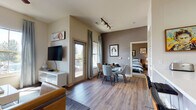 Located at 3420 S Platte River Dr in Sheridan, Colorado, this single-story residence offers a thoughtfully designed open-concept layout with contemporary finishes and a warm, inviting ambiance. The living area features rich hardwood-style flooring, vaulted ceilings, and generous natural light streaming through large windows with outdoor views. A ceiling fan, wall-mounted television, and a mix of neutral and bold accent wall tones create a comfortable, modern atmosphere. A glass door provides access to an outdoor space, further enhancing the connection to the surroundings.

The kitchen is efficiently designed with rich reddish-brown cabinetry, black appliances including a refrigerator and range, and a bright white countertop island with a single-basin sink — ideal for both meal preparation and casual dining. Adjacent to the kitchen, a compact dining area accommodates a round table with upholstered chairs, completing the open living space.

Both bedrooms offer plush carpeting, vaulted ceilings, and ample natural light. The primary bedroom includes an attached walk-in closet. The bathroom features wood-style flooring, reddish-brown vanity cabinetry, a white countertop, a large mirror, and a tub-shower combination, maintaining the consistent modern aesthetic found throughout the unit.