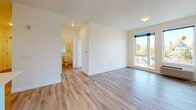 This single-floor unit is designed with a clean, contemporary aesthetic, featuring light wood-tone flooring that flows consistently throughout every room, creating a cohesive and inviting atmosphere. The kitchen is a standout feature, outfitted with flat-front light wood cabinetry, open shelving, white quartz countertops, a subway tile backsplash, and a full suite of stainless steel appliances including an electric range, over-the-range microwave, dishwasher, and a French door refrigerator. A pendant light accents the kitchen area, and a breakfast bar provides additional counter space. The open living area is bright and versatile, with white walls and large windows that invite natural light and offer views of the surrounding neighborhood greenery. The bedrooms are generously sized, each featuring the same warm wood-tone flooring, large windows for natural light, and dedicated closets with built-in rods for organized storage. The full bathroom is well-appointed with a single-sink vanity featuring warm wood cabinetry, white countertop, a large mirror, a toilet, and a shower-tub combination. The space overall presents a modern, move-in-ready environment with a neutral palette that offers flexibility for personalization.