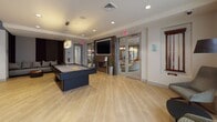 Located at 25 William T Morrissey Blvd in Boston, MA, this single-floor amenity lounge is designed for socializing, entertainment, and relaxation. The space is anchored by light hardwood flooring throughout, creating a cohesive and inviting atmosphere across its distinct zones.

The game room features a full-size pool table, a wall-mounted flat-screen TV, and a tile-surround fireplace, lending a warm and lively energy to the area. Adjacent to it, a bright lounge area is bathed in natural light from floor-to-ceiling windows dressed with sheer curtains, and includes a shuffleboard table alongside clusters of curved, modern seating.

A dedicated screening room stands out with its dramatic navy ceiling, large-format projection screen, and a generous curved sectional sofa complemented by accent chairs — ideal for movie viewings or group gatherings.

The open social lounge and dining area showcases a striking concrete communal table surrounded by contemporary mixed-style chairs, pendant lighting suspended from a dark ceiling detail, and an integrated wet bar with a white countertop. Framed artwork and carefully chosen décor add a refined, gallery-like quality throughout. The overall design blends bold color contrasts with clean, modern lines to deliver a dynamic and multifunctional community space.