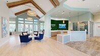Located at 7600 Heron Cir in Vero Beach, Florida, this single-story clubhouse at Aspire Vero Beach offers a well-appointed, open-concept layout designed for both community engagement and everyday comfort. The centerpiece of the space is a dramatic reception area anchored by a living moss wall bearing the Aspire Vero Beach branding, a textured herringbone-tiled front desk, and elegant pendant lighting that sets a sophisticated tone from the moment you enter. Soaring vaulted ceilings with exposed rustic wood beams define the main gathering area, which features light-toned hardwood floors, sage green accent walls, and abundant natural light streaming through oversized windows and glass doors that frame views of the surrounding Florida landscape. The living area is furnished with plush blue accent chairs and casual seating arrangements, creating an inviting atmosphere for socializing. A dedicated dining area includes a large communal table with counter-height seating and a wall-mounted display screen, ideal for group gatherings or presentations. The kitchen area offers built-in cabinetry, a compact refrigerator, and a decorative fluted backsplash. Open floating shelves styled with greenery and curated décor add warmth throughout. A separate lounge nook features large windows overlooking the pool area, completing this bright and thoughtfully designed community space.