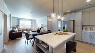 This single-story residence showcases a thoughtfully designed open-concept layout that seamlessly connects the kitchen, dining, and living areas. The kitchen is a standout feature, equipped with gray shaker-style cabinetry, stainless steel appliances, a white elongated tile backsplash, and a large central island with open lower shelving. Warm hardwood-style flooring runs throughout the kitchen and living spaces, complementing the neutral color palette. Three globe pendant lights hang above the island, adding a contemporary touch.

The living room features a textured accent wall, large windows with striped drapery that invite abundant natural light, and direct access to an outdoor area. The bedroom offers plush carpeting, a generous window framed by dark curtains, soft ambient lighting, and a calm, restful atmosphere.

A dedicated mudroom near the kitchen provides built-in shelving, hooks, and a bench for organized storage. The bathroom features light blue cabinetry with a white countertop and hardwood-style flooring. Throughout, the design balances modern finishes with warm, inviting tones, creating a cohesive and functional living environment.