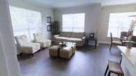 This single-floor space offers a welcoming and well-appointed interior designed for comfortable everyday living. The living area serves as the centerpiece of the home, showcasing rich, dark hardwood flooring that extends throughout and adds a warm, refined character to the space. Elegant crown molding lines the ceiling, lending a classic architectural detail that elevates the overall aesthetic.

Natural light flows generously into the living room through multiple windows dressed with crisp white horizontal blinds, creating a bright and airy atmosphere. The neutral gray walls provide a versatile backdrop that complements a range of furnishings and décor styles. The space is well-proportioned, offering ample room for comfortable seating arrangements and effortless movement throughout.

The living area is thoughtfully laid out, with enough space to accommodate a full sofa, accent chairs, and a central coffee table arrangement, making it ideal for both relaxed lounging and entertaining. A side table with a lamp adds to the functional yet stylish setup. Touches of greenery introduce a natural, lively element to the interior. Overall, the space balances classic architectural details with a clean, contemporary feel, resulting in a comfortable and inviting single-floor home.