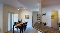 This single-floor residence is designed with an open, flowing layout that seamlessly connects the kitchen and dining areas. The dining space comfortably accommodates a full-sized table and chairs, set atop plush carpeting and bathed in natural light from a large sliding glass door that opens to an exterior patio or balcony. Vaulted ceilings amplify the sense of openness throughout, while adjustable track lighting provides focused illumination for both functional and ambient needs. A raised granite-topped kitchen counter serves as a natural divider between the cooking and dining zones, offering additional surface space and a casual breakfast bar feel. The kitchen features rich, dark granite countertops that contrast elegantly with the bright, neutral-toned walls. The overall color palette is clean and contemporary, with warm accents introduced through furnishings and décor. A hallway visible from the dining area leads to additional rooms, suggesting a well-organized floor plan. The combination of vaulted ceilings, natural light, and an open-concept design creates a welcoming and spacious ambiance throughout this single-level home.