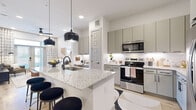 This thoughtfully designed one-bedroom, two-bathroom residence offers a contemporary open-concept layout that maximizes every square foot. The combined kitchen and living area serves as the heart of the space, anchored by a large granite-topped island with seating for multiple bar stools, illuminated by a pair of striking matte black pendant lights. The kitchen is equipped with stainless steel appliances, sage-toned cabinetry extending to the ceiling, a subway tile backsplash, and under-cabinet lighting. A built-in desk area adjacent to the kitchen provides a functional workspace.

The living room is defined by notably high ceilings, light hardwood-style flooring, a ceiling fan, and French doors with transom windows that flood the room with natural light. The primary bedroom is a comfortable retreat, featuring a window with natural light and direct access to a dedicated bathroom with granite vanity and a walk-in closet with built-in rod storage. A second full bathroom features a soaking tub enclosed in floor-to-ceiling neutral tile with matte black fixtures. The overall aesthetic throughout is clean, modern, and cohesive.