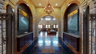 This single-story facility showcases a striking rustic design rooted in Texas Hill Country aesthetics. The grand entrance corridor is defined by dramatic limestone archways, polished slate-and-stained concrete flooring, and wrought-iron chandeliers suspended from soaring ceilings. Large-scale artwork flanks the entryway, while dark wood cabinetry with granite countertops lines both sides, establishing a formal yet welcoming reception area.

Beyond the entry arch, a lounge area features natural light from multiple wood-framed windows and French doors, stone wainscoting, and comfortable seating arranged around a round stone-top coffee table. A dedicated office area includes a substantial executive desk, stone column accents, and polished concrete floors that continue throughout.

The game room offers vaulted ceilings with exposed wood beams, a full pool table, a shuffleboard table, and multiple wall-mounted televisions. An adjacent lounge nook with turquoise bistro tables and leather barrel chairs serves as a social gathering spot. A fully equipped kitchenette features dark wood cabinetry, granite countertops, stainless steel appliances, and a coffee station. A compact business center with faux-finished walls provides workstations for residents. Throughout, the warm stained concrete floors, limestone detailing, and iron fixtures create a cohesive, upscale rustic ambiance.