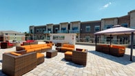 This outdoor terrace is a well-appointed communal space designed for relaxation and social gatherings. The patio is finished with large-format stone pavers that lend a clean, structured aesthetic underfoot, while providing ample room for multiple seating arrangements. Generously sized wicker lounge sets with bold orange cushions are arranged throughout the space, offering comfortable spots for conversation and leisure. A large patio umbrella on one end provides shade on sunny days, adding practical comfort to the open-air setting.

The terrace faces a contemporary multi-story building clad in a combination of dark gray horizontal siding and warm brick masonry. The building features glass-panel balcony railings on the upper level, contributing to a modern architectural character. A central utility or service structure sits on the terrace level, subtly dividing zones within the space without disrupting the open feel.

The setting benefits from abundant natural light and open sky views, creating a bright and inviting atmosphere. Overall, the terrace presents a polished, resort-inspired outdoor environment well-suited for communal use.