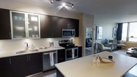 This single-floor residence showcases a thoughtfully designed open-concept layout that seamlessly connects the kitchen and living areas. The kitchen is a standout feature, outfitted with rich dark cabinetry that contrasts elegantly against crisp white countertops, including a spacious island that serves as both a prep surface and casual dining area. Stainless steel appliances — including a range with oven, over-the-range microwave, and dishwasher — lend a polished, modern aesthetic throughout. Upper cabinets feature frosted glass panel inserts, adding a refined touch to the contemporary design. Track lighting overhead provides focused illumination across the workspace, enhancing both function and ambiance. The kitchen flows naturally into a bright and comfortable living area, visible in the background, which is furnished with modern seating and accented with tasteful décor. The open sightlines between rooms create a sense of spaciousness and connectivity throughout the home. Dark hardwood-style flooring runs throughout, grounding the space with warmth and continuity. The overall design balances clean lines, neutral tones, and modern finishes to create a stylish and highly functional living environment.