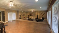 This single-floor space makes a strong impression with its open, airy layout and warm, inviting aesthetic. The main living area is expansive, showcasing rich hardwood flooring that extends throughout and contributes to a cohesive, grounded feel. Neutral taupe walls provide a versatile backdrop that complements both the flooring and the thoughtfully arranged furnishings.

The living area is anchored by a classic seating arrangement, including a plush sofa, accent chairs with decorative pillows, and a central coffee table atop a area rug — creating a defined and welcoming gathering space. A sleek black chaise lounge adds a bold, modern contrast near the seating area. Decorative wall art and sculptural accents add personality and visual interest throughout.

Overhead, a ceiling fan with light fixture serves as a functional centerpiece, complemented by recessed lighting that provides even, well-distributed illumination. Crown molding runs along the upper perimeter of the walls, adding an elegant architectural detail. A large window along one wall allows natural light to filter in. The overall ambiance is warm, polished, and comfortably livable — a well-proportioned space that balances style and everyday functionality with ease.