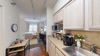 Located at 635 Cotanche St in Greenville, NC, this one-bedroom, one-bathroom unit offers a thoughtfully organized single-floor layout designed to maximize every square foot. The space is divided into distinct functional areas that flow seamlessly from one to the next.

The galley-style kitchen is positioned at the entry and features light wood cabinetry, a continuous countertop with a stainless steel sink, a gas range, and an over-the-counter microwave — providing efficient meal preparation within a compact footprint. Hardwood flooring runs through the kitchen and living corridor, lending warmth to the space.

The open living area transitions directly from the kitchen and offers room for a sofa, a dedicated workspace with a desk and chair, and wall-mounted décor. Exposed ceiling conduit adds an industrial character, complemented by soft ambient lighting from floor lamps. A window at the far end allows natural light to brighten the living and sleeping areas.

The bedroom is generously proportioned relative to the overall unit size, featuring carpeted flooring, neutral walls, and ample room for a full-sized bed with accompanying furniture. The bathroom is clean and functional, with a vanity, mirror, toilet, and towel fixtures in a compact, well-appointed layout.