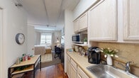 Located at 635 Cotanche St in Greenville, NC, this one-bedroom, one-bathroom unit offers a thoughtfully organized single-floor layout designed to maximize every square foot. The space is divided into distinct functional areas that flow seamlessly from one to the next.

The galley-style kitchen is positioned at the entry and features light wood cabinetry, a continuous countertop with a stainless steel sink, a gas range, and an over-the-counter microwave — providing efficient meal preparation within a compact footprint. Hardwood flooring runs through the kitchen and living corridor, lending warmth to the space.

The open living area transitions directly from the kitchen and offers room for a sofa, a dedicated workspace with a desk and chair, and wall-mounted décor. Exposed ceiling conduit adds an industrial character, complemented by soft ambient lighting from floor lamps. A window at the far end allows natural light to brighten the living and sleeping areas.

The bedroom is generously proportioned relative to the overall unit size, featuring carpeted flooring, neutral walls, and ample room for a full-sized bed with accompanying furniture. The bathroom is clean and functional, with a vanity, mirror, toilet, and towel fixtures in a compact, well-appointed layout.