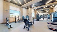 Located in Maricopa, Arizona, this contemporary fitness center offers a thoughtfully designed exercise space that combines functionality with modern aesthetics. The facility occupies a single floor and provides ample room for a comprehensive workout experience.

The exercise room features soaring ceilings with a coffered design accented by sleek linear LED lighting, creating a bright and energizing atmosphere. Warm wood-toned wall panels add a refined touch to the otherwise clean, neutral palette of white walls and light-colored flooring. Large windows along multiple walls invite abundant natural light and frame views of the surrounding Arizona landscape, including palm trees and outdoor amenity areas.

The space is outfitted with a diverse array of equipment to support both cardiovascular and strength training. Multiple treadmills are arranged along the windowed wall, alongside a rowing machine and a stair climber for cardio variety. The strength training area includes selectorized cable and weight machines, adjustable benches, a barbell and weight rack, and additional resistance equipment. Floor-to-ceiling glass partition doors lead to a connected section of the facility, expanding the workout area and enhancing the open, airy feel. Full-length mirrors further amplify the sense of space. The combination of modern design, natural light, and comprehensive equipment makes this fitness center a well-appointed destination for residents in the Maricopa community.