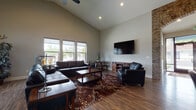 This single-story space combines rustic and modern design elements to create a welcoming, functional environment. The entry welcomes visitors through glass-paneled doors into an open area with soaring vaulted ceilings and rich hardwood-style flooring that flows throughout. A decorative exposed brick archway serves as a prominent architectural feature, framing the transition between areas and adding warmth and character to the space.

The main lounge area is generously proportioned, featuring a row of large windows that flood the room with natural light and offer views of the surrounding neighborhood. Dark leather seating, a tufted sofa, an ornate area rug, and a wall-mounted flat-screen television create a comfortable and well-appointed gathering space. A ceiling fan enhances the airy feel of the high vaulted ceilings.

An adjacent office or meeting area features a commanding floor-to-ceiling stacked stone fireplace with a rustic wood mantle, serving as the room's focal point. A centrally placed desk and open floor plan make this area well-suited for professional use. Natural light, neutral wall tones, and consistent flooring unify the overall ambiance, resulting in a polished yet inviting atmosphere throughout.