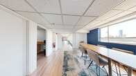 This well-appointed single-floor communal space offers an impressive layout designed for both collaboration and relaxation. Light hardwood flooring flows throughout the interior, complementing the acoustic tile ceilings fitted with recessed lighting. A wall of floor-to-ceiling windows runs along one side of the main area, flooding the space with natural light and framing sweeping views of the Boston skyline and waterfront — a defining architectural feature of the space.

The open main area is thoughtfully arranged with multiple dining and lounge zones, featuring modern wire-frame chairs, casual tables, a foosball table, and comfortable seating, all set against soft teal-painted walls accented with curated artwork. A long communal work table with bar-height seating and a navy accent wall anchors a more focused workspace toward one end of the floor.

A compact kitchenette provides functional support with ample counter space, a range, refrigerator, and sink — ideal for catering or everyday use. French doors lead directly to a spacious outdoor terrace adorned with string lights, offering additional seating and open-air panoramic views of the surrounding cityscape on multiple sides. The overall ambiance is bright, modern, and inviting, making the space well-suited for gatherings, meetings, or everyday communal use.