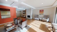This single-floor communal lounge is designed with a refined yet welcoming aesthetic, centered around a striking floor-to-ceiling stacked-stone fireplace that serves as the architectural focal point of the space. The fireplace divides the open layout into distinct seating zones, each thoughtfully furnished to encourage conversation and relaxation.

Light hardwood flooring runs throughout, complementing the neutral warm tones of the walls and tray ceilings, which add architectural depth and a polished finish. Recessed lighting is embedded throughout the ceiling, supplemented generously by natural light streaming through expansive floor-to-ceiling glass sliding doors along one wall. These doors open to a landscaped outdoor area, framing views of colorful seasonal foliage and providing a seamless indoor-outdoor connection.

Seating areas are furnished with upholstered armchairs and accent pieces in coordinating gray and orange tones, grounded by area rugs. An accent wall painted in warm terracotta adds a bold visual contrast, paired with curated artwork and decorative accessories throughout. A secondary seating nook offers a quieter retreat with patterned accent chairs and a small side table. The overall ambiance is contemporary and inviting, balancing comfort with stylish design elements.
