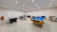 This well-appointed game room offers a generous, open layout designed entirely for recreation and socializing. The space is finished with smooth concrete-style flooring and subtly textured neutral walls that create a clean, modern backdrop throughout. A drop ceiling with a grid of recessed LED downlights provides bright, even illumination across the entire room, ensuring excellent visibility for gameplay.

The room is equipped with three full-sized game tables: a classic wood-framed pool table with a blue felt surface, a regulation-sized ping pong table, and a sleek air hockey table. These are arranged with ample clearance between them, allowing multiple games to be played simultaneously without interference. A wall-mounted flat-screen television is prominently positioned on one wall, adding a media viewing element to the space.

A comfortable lounge corner features a sofa, armchairs, and a bistro-style high-top table with bar chairs, offering a relaxed area for guests to unwind between games. Framed coastal artwork adorns the walls, lending a light, welcoming aesthetic. A wall-mounted mini-split air conditioning unit ensures year-round climate comfort, making this a functional and inviting entertainment destination.