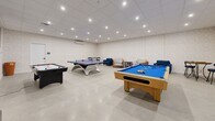 This well-appointed game room offers a generous, open layout designed entirely for recreation and socializing. The space is finished with smooth concrete-style flooring and subtly textured neutral walls that create a clean, modern backdrop throughout. A drop ceiling with a grid of recessed LED downlights provides bright, even illumination across the entire room, ensuring excellent visibility for gameplay.

The room is equipped with three full-sized game tables: a classic wood-framed pool table with a blue felt surface, a regulation-sized ping pong table, and a sleek air hockey table. These are arranged with ample clearance between them, allowing multiple games to be played simultaneously without interference. A wall-mounted flat-screen television is prominently positioned on one wall, adding a media viewing element to the space.

A comfortable lounge corner features a sofa, armchairs, and a bistro-style high-top table with bar chairs, offering a relaxed area for guests to unwind between games. Framed coastal artwork adorns the walls, lending a light, welcoming aesthetic. A wall-mounted mini-split air conditioning unit ensures year-round climate comfort, making this a functional and inviting entertainment destination.