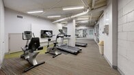 Located at 6030 Bathurst Street in Toronto, Ontario, this fitness facility offers a dedicated space for cardiovascular exercise and general wellness. The room is laid out in an elongated floor plan, providing a functional and organized workout environment across a single level.

The space is equipped with multiple cardio machines, including Vision T80 treadmills and upright and recumbent exercise bikes, all positioned on protective rubber mats atop a multi-toned, patterned carpet. A wall-mounted flat-screen television is installed for entertainment during workouts. Large mirrors line one wall, visually expanding the space and allowing users to monitor their form.

Overhead, exposed pipes and structural elements run across the ceiling, lending the space an industrial character. Fluorescent strip lighting provides consistent, bright illumination throughout. The white-painted concrete block walls reinforce the utilitarian aesthetic of the facility.

A glass-partitioned room is accessible from the main fitness area, visible through full-height aluminum-framed glass panels, offering an additional separate space. Emergency exit signage and safety features are clearly marked throughout.

The layout ensures easy circulation between equipment stations, making this a practical and well-appointed fitness amenity suited for regular use.