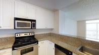 This single-floor space showcases a well-appointed kitchen as its focal point. The kitchen features granite countertops with warm golden and brown tones, providing both durability and visual appeal. Crisp white cabinetry lines the walls, offering generous upper and lower storage with simple, classic hardware. The layout follows an L-shaped configuration, maximizing counter space and workflow efficiency. Black appliances, including an electric range, built-in microwave, and dishwasher, contrast cleanly against the white cabinetry and granite surfaces. The textured ceiling adds subtle dimension to the space, while neutral-toned walls contribute to a bright, clean aesthetic. The kitchen opens toward an adjacent area, suggesting a connected, open feel to the overall floor plan. The overall ambiance is practical and tidy, with a timeless color palette of white, black, and warm stone tones that creates a cohesive and inviting environment suited for everyday use.