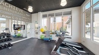 This contemporary fitness center is designed to inspire and energize, featuring a single open-plan layout that maximizes functionality and flow. The space is framed by expansive floor-to-ceiling windows on multiple walls, flooding the interior with natural light and offering broad outdoor views, including scenic waterfront vistas and palm tree-lined surroundings. The dark, drop-panel ceiling provides a striking contrast to the bright, white walls and creates a sleek, modern aesthetic. Durable rubber flooring covers the entire workout area, offering both safety and comfort underfoot. The cardio zone is well-stocked with treadmills and stationary spin bikes, while the strength training area includes a multi-functional cable machine and a full rack of dumbbells. Functional training is supported by a dedicated station with medicine balls, resistance bands, and stability balls. A large mirrored wall reflects the space and enhances the sense of openness, accompanied by mounted flat-screen TVs for entertainment during workouts. Adding a motivational touch, the walls are adorned with bold typographic murals featuring fitness-inspired words such as "Achieve," "Discipline," "Endurance," and "Inspire," setting an energetic and goal-driven atmosphere throughout the facility.