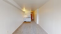 This single-floor unit offers a straightforward, open layout designed for efficient use of space. The main living area is carpeted in neutral tones and features white walls with a flat ceiling accented by a structural beam detail. A window along one wall allows natural light to enter and houses a wall-mounted air conditioning unit, complemented by a baseboard heating system for year-round comfort.

The kitchenette is positioned at one end of the main space and transitions to tile flooring. It is outfitted with white cabinetry above and below a granite-patterned countertop that integrates a stainless steel sink. A white electric coil range and a full-size white refrigerator complete the appliance package. An under-cabinet range hood provides ventilation, and a wall sconce adds focused lighting to the cooking area.

The bathroom is accessible via a wood-panel door and features a white vanity with ample counter space, a toilet, and a tiled walk-in shower. The floor is finished with a mosaic tile pattern in soft gold tones. A louvered window provides ventilation, and clean white walls throughout contribute to the bathroom's bright, functional atmosphere. Overall, the unit presents a clean and practical living environment.